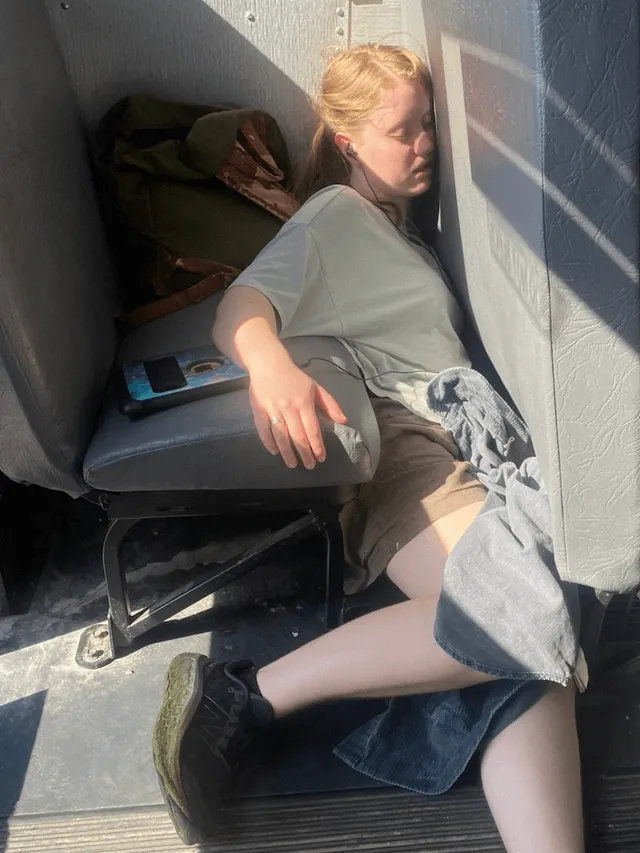 A girl asleep on a bus, wedged into the corner, lit by diagonal afternoon light